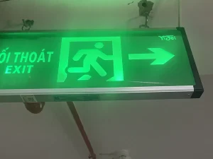 Đèn chỉ dẫn lối thoát nạn EXIT màu xanh lá có hình người chạy và mũi tên chỉ hướng, treo trên trần hành lang