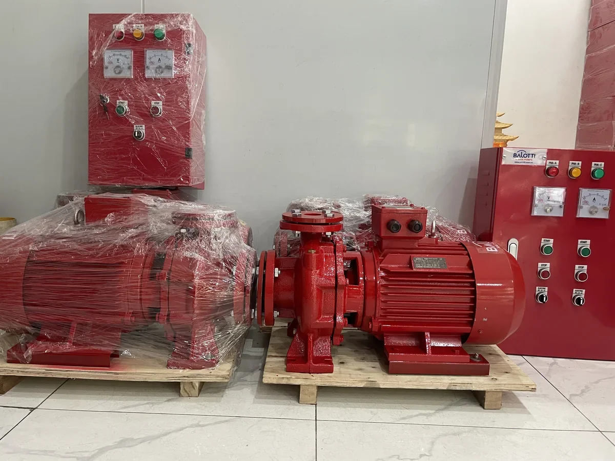 Hai máy bơm chữa cháy trục ngang sơn đỏ còn bọc màng nilon đặt trên pallet gỗ, phía sau là hai tủ điều khiển bơm có đồng hồ và nút bấm