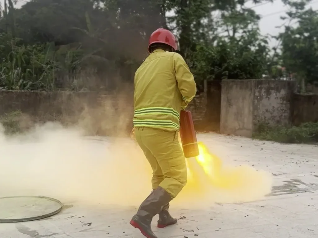 Người mặc quần áo chữa cháy, đội mũ đỏ dùng bình chữa cháy bột xách tay phun dập lửa khay trong buổi thực tập phương án ngoài trời