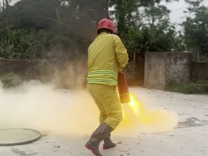 Người mặc quần áo chữa cháy, đội mũ đỏ dùng bình chữa cháy bột xách tay phun dập lửa khay trong buổi thực tập phương án ngoài trời
